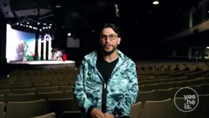 Man in a blue patterned jacket stands in an empty auditorium, showcasing a stage setup in the background.