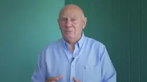 Elderly man in a light blue shirt speaking against a green background, conveying an engaging message or story.