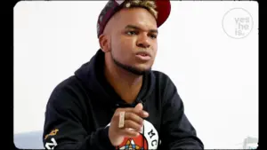 Young man in a black hoodie and cap, passionately speaking during a discussion or presentation. Engaged and expressive demeanor.