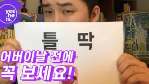 Person holding a sign with text in Korean, promoting content to view before a specific event, captured in a casual setting.