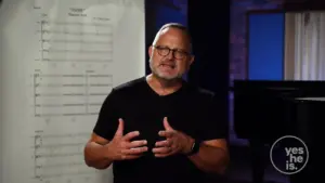 Man in a black shirt gestures while explaining concepts, with a chart and piano in the background during a presentation.