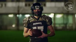 Football player in a black camouflage uniform, holding a ball, standing on a field with a focused expression.