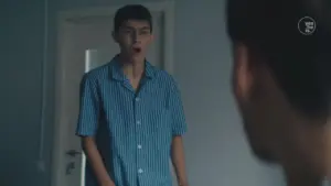 Teen in striped pajamas stands surprised in a doorway, facing a person just out of frame. Soft lighting enhances the scene.