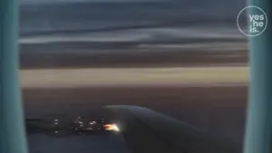 View from an airplane window at sunset, showcasing wings and distant city lights against a dramatic sky.