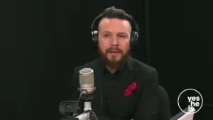 Man with a beard wearing a black shirt and headphones, sitting in front of a microphone in a studio setting.