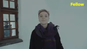 Smiling woman in a cozy indoor setting, wearing a purple scarf and standing against a light wall.