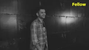 Smiling man in a plaid shirt standing against a textured black wall with dramatic lighting, showcasing a relaxed vibe.