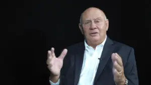 Older man in a suit gesturing while seated against a black background, conveying a sense of authority and engagement.