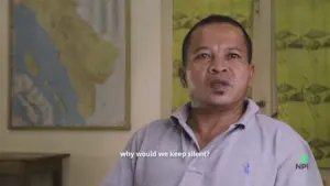 Man in a gray polo shirt speaks in front of a map, questioning silence on important issues.