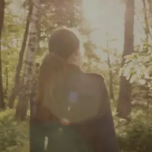 Woman walking through a sunlit forest, surrounded by lush greenery and trees, capturing a serene outdoor moment.