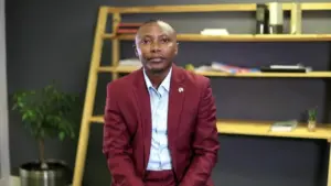 Professional man in a red suit seated in a modern office space with bookshelves and a plant in the background.