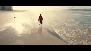 Child walking along the shoreline with gentle waves crashing, sunlight reflecting on wet sand.