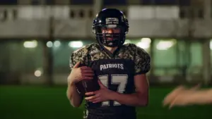 A football player in a camouflage jersey holds a ball, standing on a well-lit field at night.