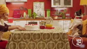 A cozy kitchen scene with a woman and a dog at a table, featuring a cake and fresh juice, creating a warm atmosphere.