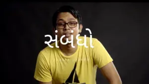 Young man in a yellow shirt with glasses, smiling, against a dark background featuring the word "संवंध" in white.