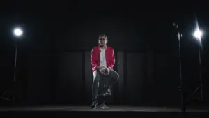 Man in a red jacket sitting on a stool under spotlight, creating a dramatic atmosphere in a dark room.