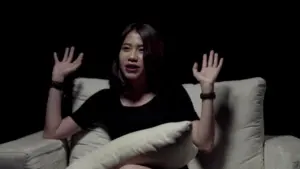 Young woman sitting on a couch with hands raised, against a black background, conveying surprise or excitement.