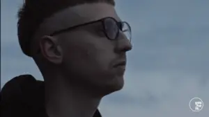 Profile of a young man with glasses outdoors, looking contemplatively against a cloudy sky backdrop.