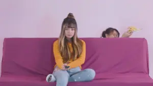 Girl sitting on a pink couch watching TV, while another child playfully peeks from behind with a banana.