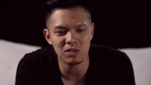 Close-up of a young man with short hair, wearing a black shirt, expressing deep emotions against a dark background.