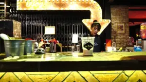 Dimly lit bar scene with a bartender, glowing arrow sign, and a foreground of drinks on a vibrant, textured countertop.