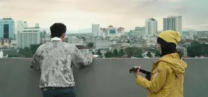 Couple overlooking city skyline at sunset, with one holding a guitar and the other in a yellow jacket.