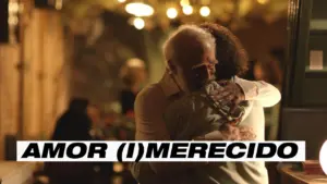 Elderly couple embracing in a warm, dimly lit restaurant, conveying love and connection. Text overlay: "AMOR (I)MERECIDO."