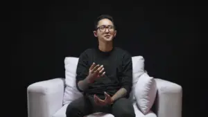 Person sitting on a white couch against a dark background, engaging with the audience and expressing ideas.