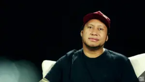 Man in a black t-shirt and red cap sitting on a couch against a dark background, conveying a thoughtful expression.