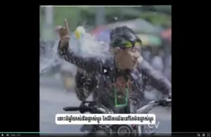 A cyclist splashes through water, joyfully gesturing with one hand during a vibrant outdoor event.