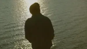 Silhouette of a person standing by the shimmering water, reflecting sunlight at sunset, creating a serene atmosphere.