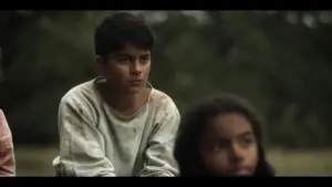 Two children, one boy with a serious expression and a girl in the background, sit outdoors in a natural setting.