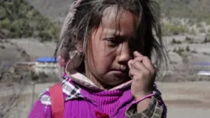 Young girl with a solemn expression wearing a purple sweater and a plaid shirt, set against a blurred mountainous background.