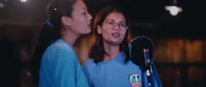 Two women singing in a recording studio, wearing matching blue shirts, with a microphone and soft lighting in the background.