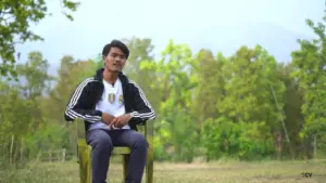 Young man sitting on a chair in a lush green outdoor setting, wearing a sporty jacket, amidst trees and mountains.