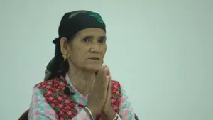 Elderly woman in traditional dress with headscarf, hands clasped together in prayer, expressing devotion and spirituality.