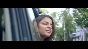 Young woman enjoying a breeze while leaning out of a car window, with trees and a street visible in the background.