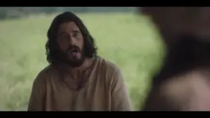 Man with long hair and beard speaking in a natural outdoor setting, highlighting a moment of dialogue.