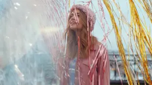 Woman in pink hat and jacket stands under colorful streamers, smiling and enjoying a festive outdoor atmosphere.