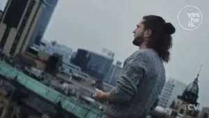 Man with long hair in a bun stands on a rooftop, overlooking a city skyline with modern buildings under a cloudy sky.