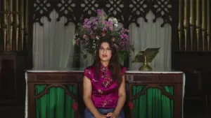 Woman in a pink blouse seated in a floral-decorated setting with a wooden backdrop and decorative elements.