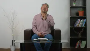 Man in a pink shirt sitting on a brown sofa, gesturing while facing the camera, with a bookshelf in the background.