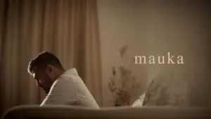 Man sitting pensively on a couch in a softly lit room, with the word "mauka" visible beside him.