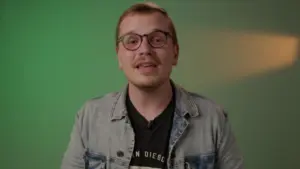 Person with glasses wearing a denim jacket, smiling in front of a green background, promoting a positive vibe.