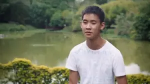 Teen boy in a white t-shirt sitting near a serene lake surrounded by lush greenery, exuding a calm atmosphere.