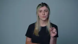 Woman with long blonde hair, wearing a black shirt, gesturing thoughtfully against a simple gray background.