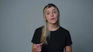 Woman with blonde hair wearing a black shirt, speaking confidently against a plain gray background.