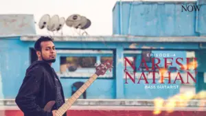 Musician Naresh Nathan poses with a bass guitar on a colorful rooftop, surrounded by urban elements and satellite dishes.