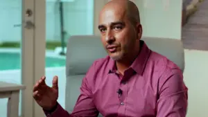 Bald man in a pink shirt discussing ideas, seated in a modern room with a pool view in the background.