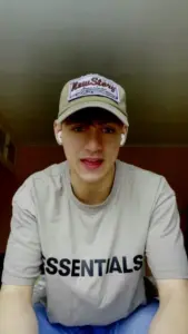 Young man wearing a khaki cap and "ESSENTIALS" t-shirt, smiling in a brightly lit room.